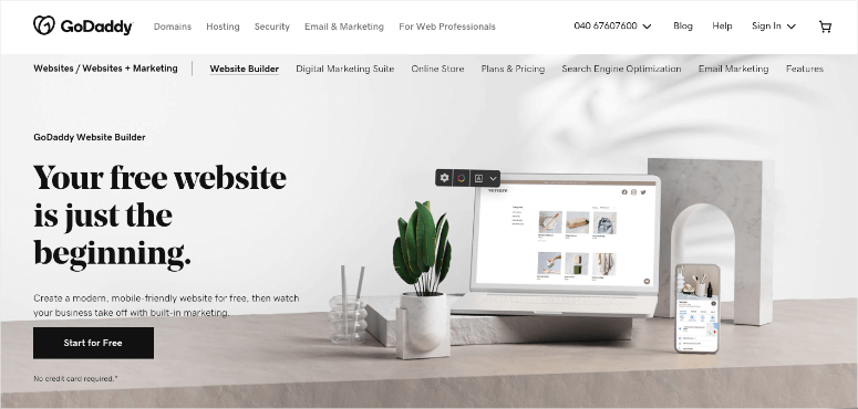 GoDaddy website builder ad with a laptop and phone displaying a website, set on a desk with plants and decor. Text: "Your free website is just the beginning."