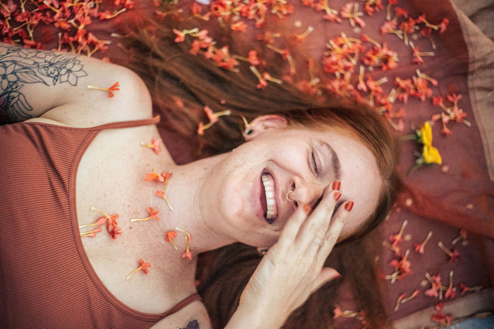A red-haired woman lying on a terracotta-colored surface scattered with small orange and yellow flowers, laughing with her eyes closed, hand resting on her cheek, floral tattoo visible on her upper arm.
