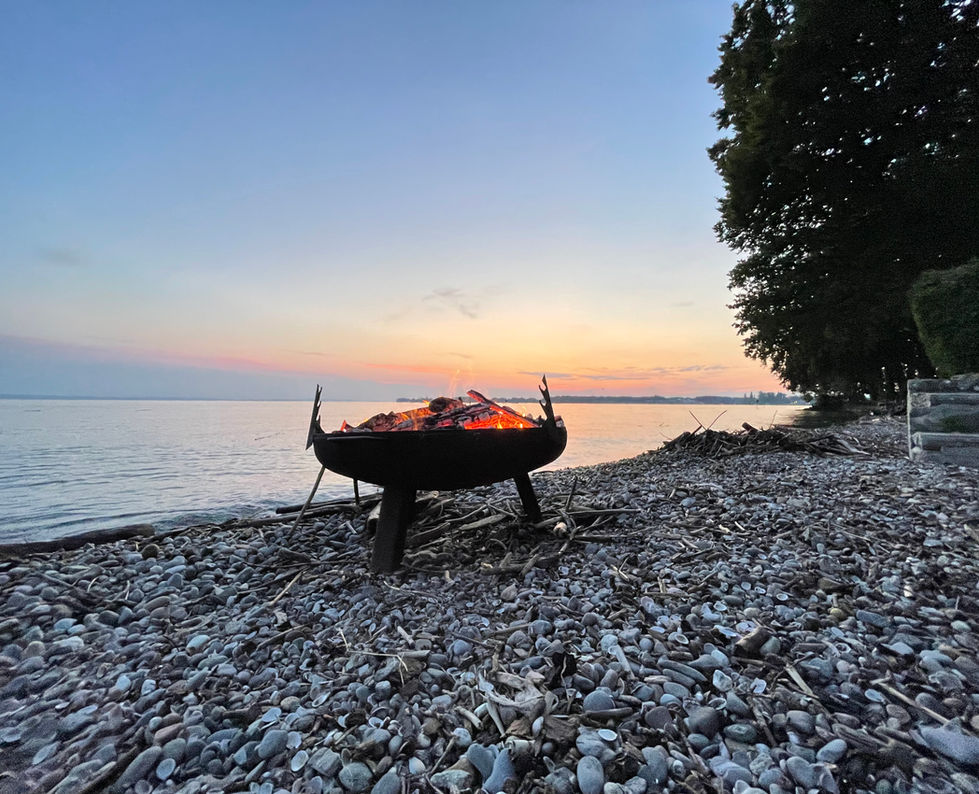 Immagine della spiaggia privata di Villa Noho sul Lago di Costanza con un focolare. Il focolare è in metallo e ha un diametro di circa un metro. Viene alimentato a legna e diffonde una luce calda e accogliente al tramonto. Sullo sfondo si vede il Lago di Costanza.