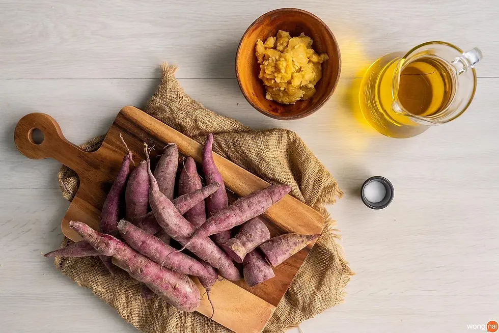 Purple sweet potatoes on a wooden board with a bowl of mashed potatoes and a glass jug of oil, set on a burlap cloth background.