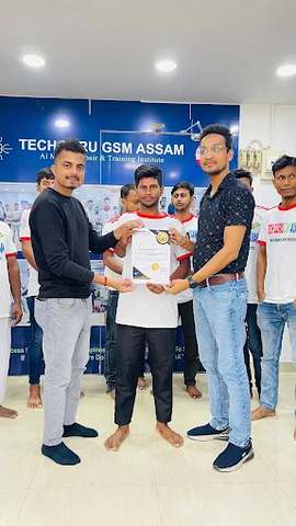 Two instructors from TechGuru Assam presenting an NSDC-approved certificate to a male student in a white "TECH GURU ASSAM" t-shirt, while other students look on.