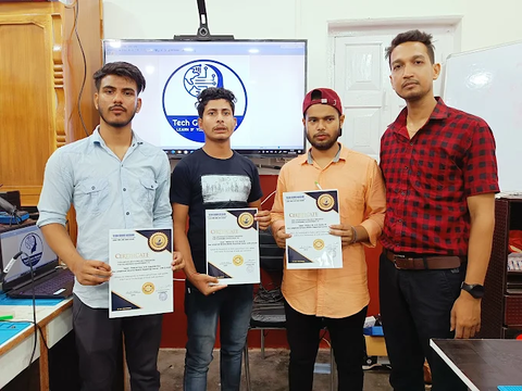 Three male students at TechGuru Assam, each holding their newly acquired NSDC-approved certificates, with a laptop and a large screen displaying the TechGuru logo in the background.