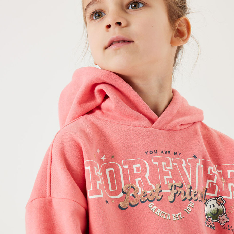 Junges Mädchen trägt rosa Kapuzenpullover mit glitzerndem Schriftzug.