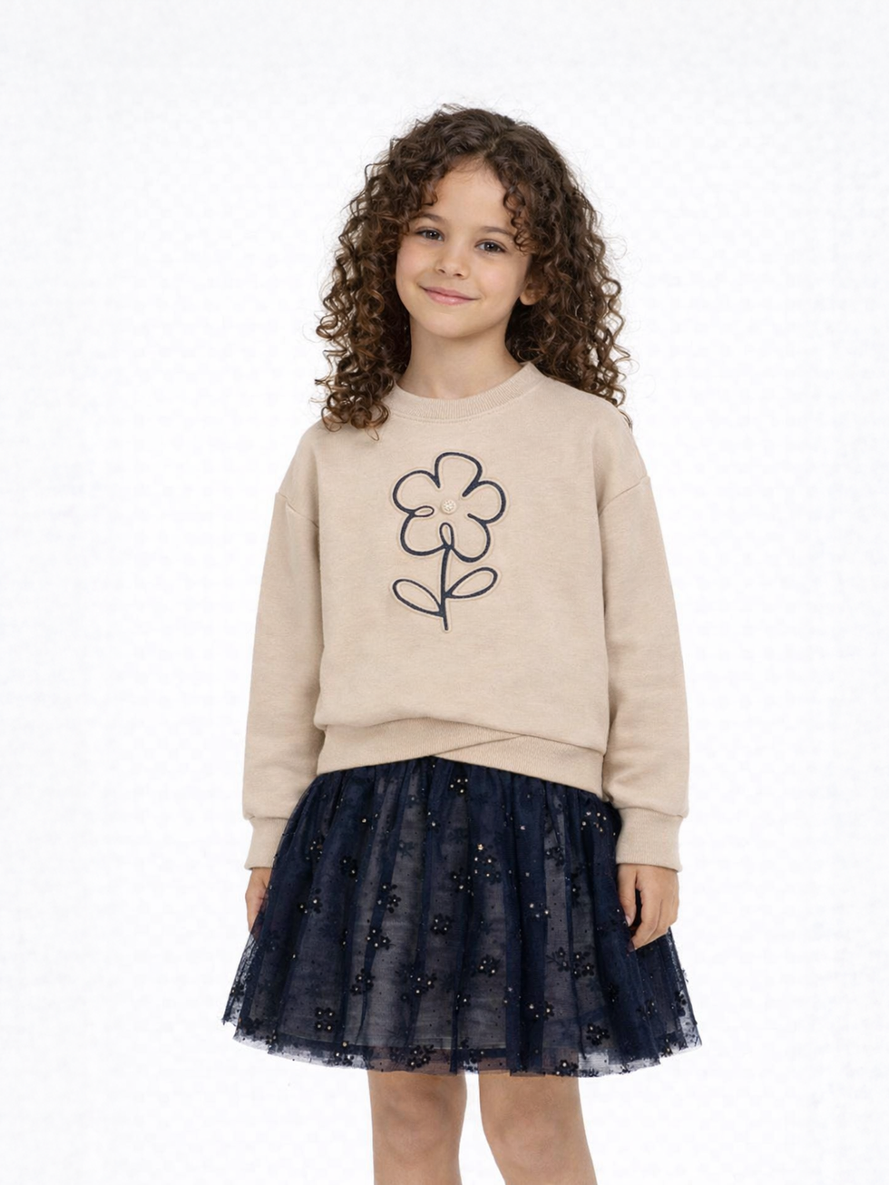 Lächelndes Mädchen in beigem Sweatshirt mit Blumenprint und blauem Tüllrock.