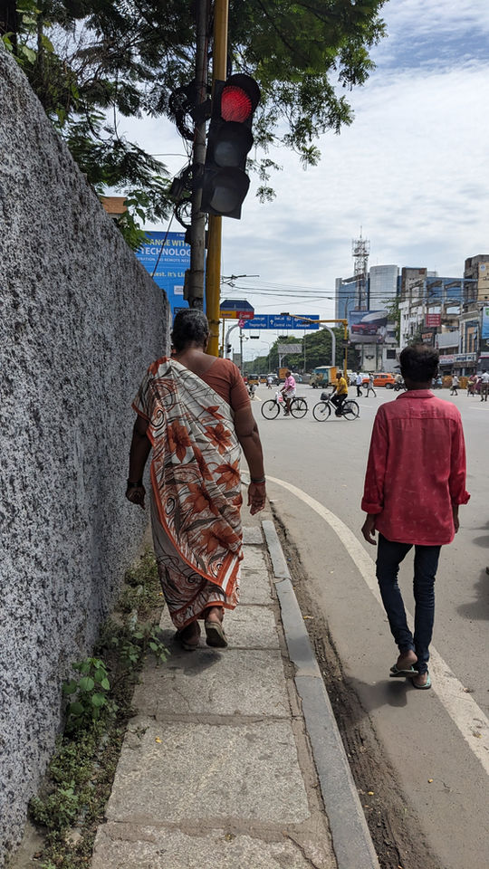Two women walk beside each other, one on a city sidewalk and the other on the carriageway.
