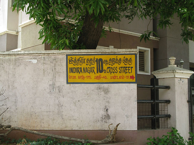 Yellow street sign: Indira Nagar 10th Cross Street written in English and Tamil on a white wall, along with red arrows. 