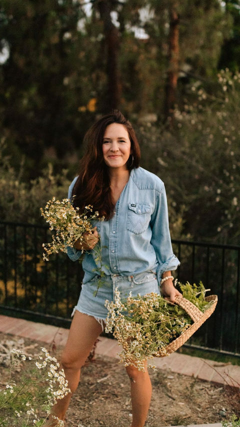 Bailey Van Tassel holding baskets of flowers, standing outside in a garden, wearing a denim shirt and shorts.