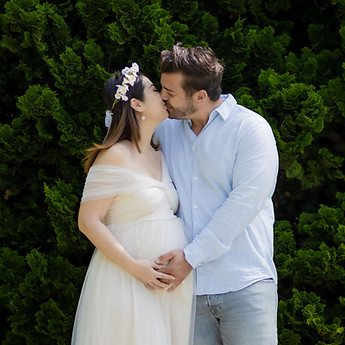 The father-to-be kisses the mother-to-be on the forehead, in front of a hedge