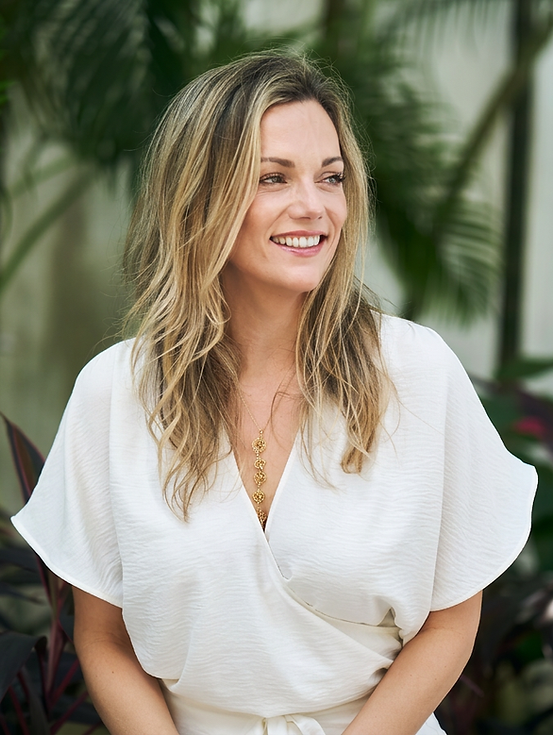 Studio photography of a smiling woman in a white ensemble, set against lush green foliage. The scene radiates a warm, natural, and friendly atmosphere.