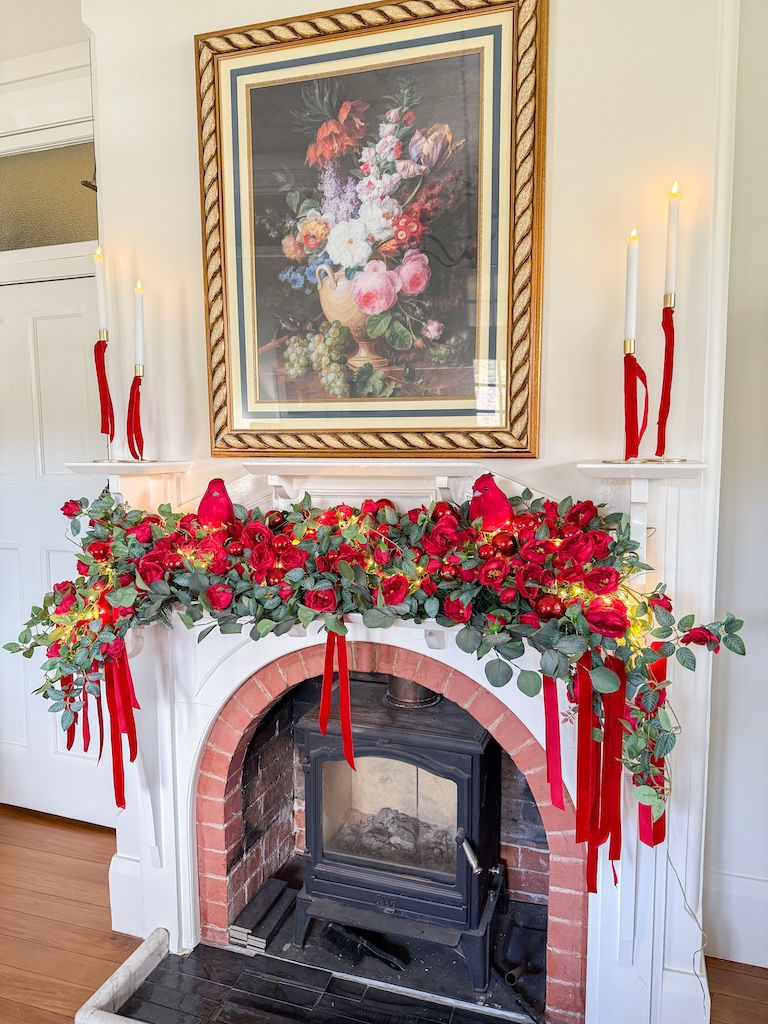 Floral mantel arrangement with red roses, greenery, and red ribbons above a fireplace. Lit candles and a framed flower painting above.