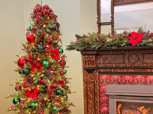 Traditional Christmas tree decorated with red and green ornaments, ribbon and natural textures beside a festive mantel, reflecting classic Christmas colour trends for 2025.