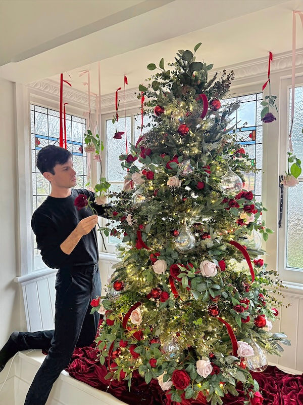 A person decorates a festive tree adorned with red and pink roses, glass baubles, and lights. Bright room with a white and red theme.
