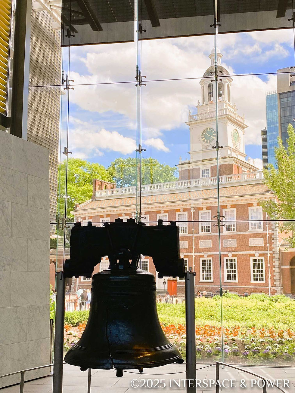 The Liberty Bell indoors against a glass wall, with Independence Hall visible outside; clear sky, lush greenery, and flowers around.