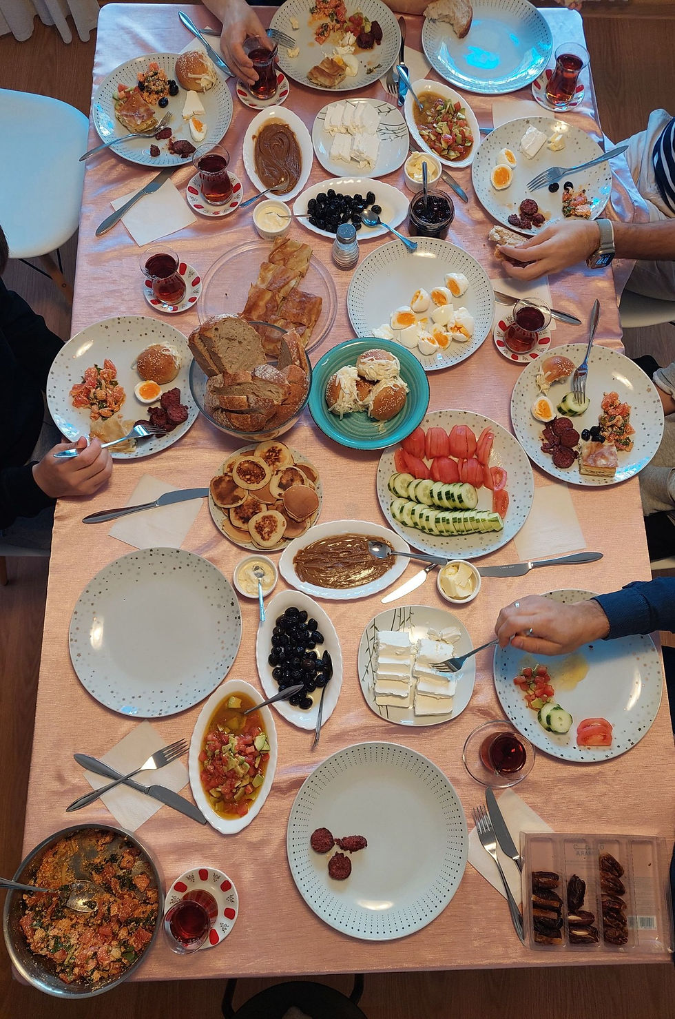 Overhead view of a group meal with plates of bread, cheese, eggs, tomatoes, olives and tea on a pink tablecloth, hands serving.