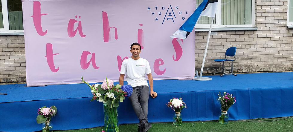 Smiling man sits on a blue stage before a pink banner reading tähis, with flower bouquets and an Estonian-style flag nearby.