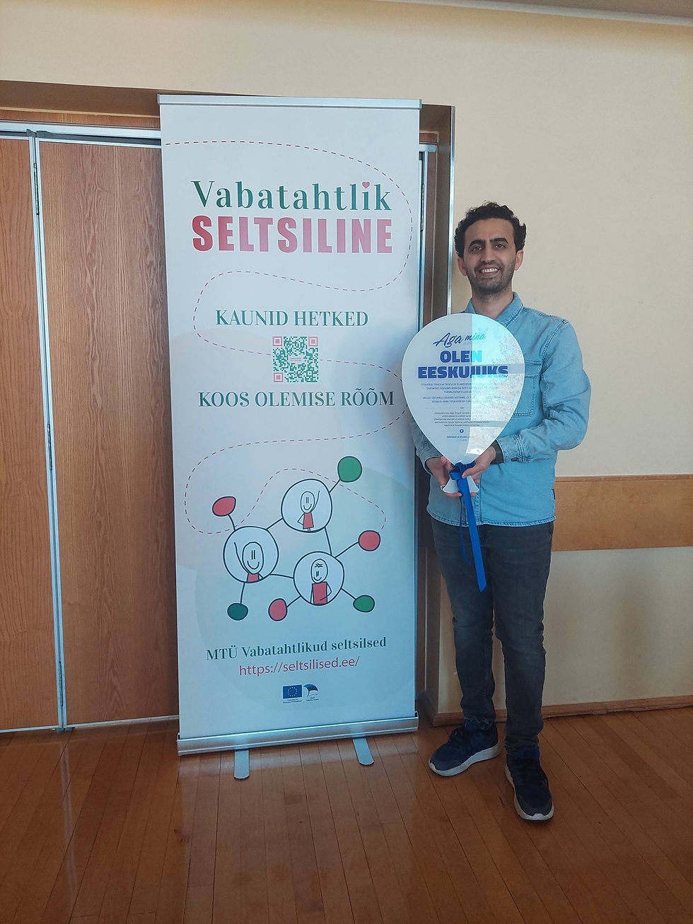 Smiling man holds a balloon-shaped sign beside a banner reading Vabatahtlik SELTSILINE, Kaunid hetked, koos olemise rõõm.
