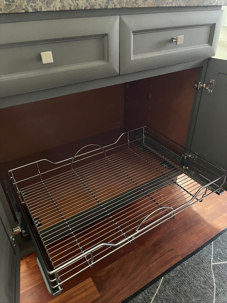 Open gray kitchen cabinet with a metal pull-out rack, set on a wooden floor. Marble countertop visible above. No text or people present.