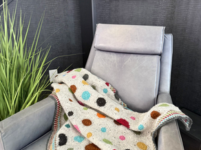 Crochet blanket with colorful polka dots draped over a gray chair. Background is dark with textured patterns, creating a cozy vibe.