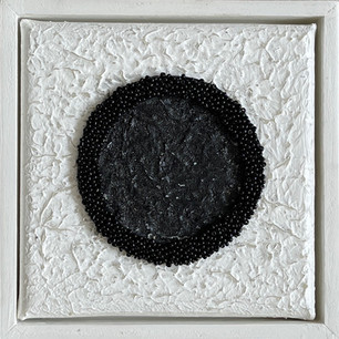 A black circle made of beads and black paint on a white textured background in a frame