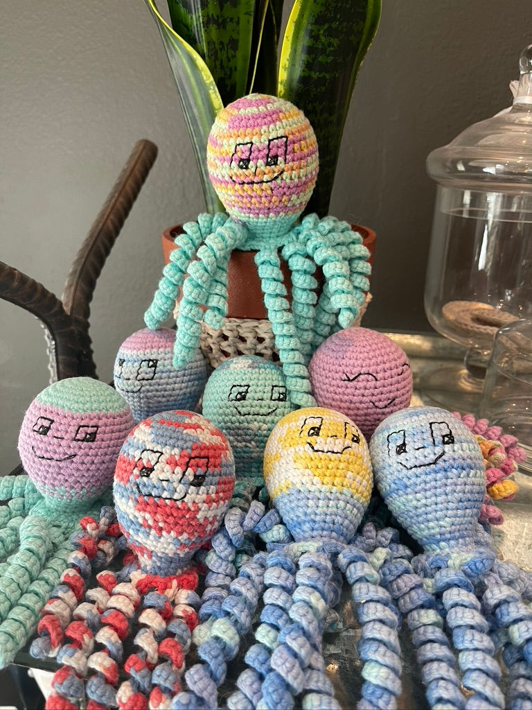 Colorful crocheted octopus toys with smiling faces on a table; a plant and glass jar are in the background, adding a playful mood.