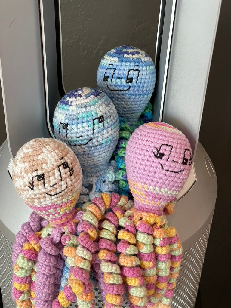 Crocheted octopus toys with smiling faces in pastel colors sit in a silver container. The mood is cheerful and playful.