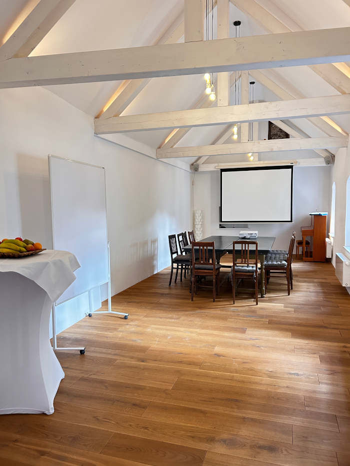 Seminarraum mit offener, weißer Balkendecke, historischen Tischen und Stühlen, eine Projektionsfläche und ein Whiteboard