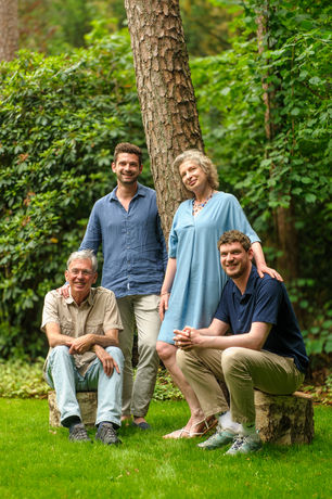 zoon en moeder staan tegen boom in tuin, vader en andere zoon zitten en kijken lachend naar camera