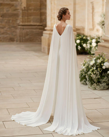Robe de mariée - combinaison pantalon Haela signée Alma Novia, style aérien avec sa cape fluide, son décolleté en V, à découvrir dans la boutique Noces de Cristal à Nancy.