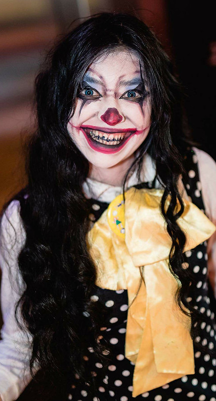 Closeup image of smiling woman dressed as a clown with white face paint, a red nose, blue eyeshadow, and painted red lips. Her teeth are painted black.