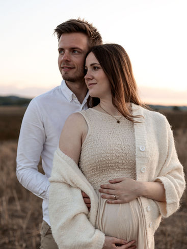 schwangerschaft. eine schwangere frau mit beigem kleid und strickjacke - dahinter steht ihr freund mit weißem hemd. ein schangerschaftsbild der beiden auf einer braunen wiese