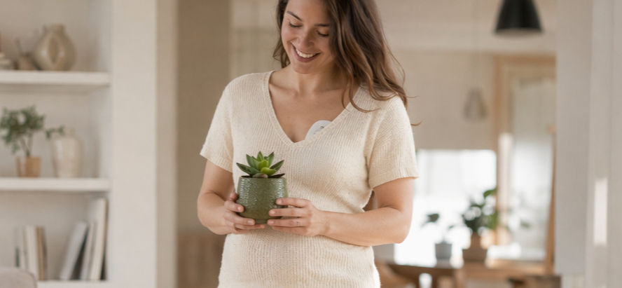 Mamá joven y feliz caminando en un living room moderno y aesthetic, utilizando de forma discreta un extractor de leche manos libres bajo su ropa para su lactancia, mientras sujeta una planta.