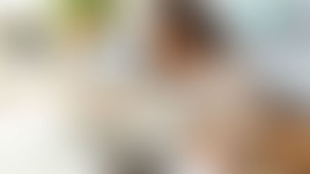 Woman in white robe sits on bedroom floor, using a laptop and taking notes. Bedroom setting with bed, plants, and soft lighting. Relaxed mood.