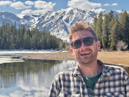 A man wearing a flannel collared shirt in front of gorgeous, snow-capped mountains 
