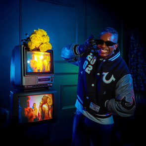 Man in a varsity jacket poses playfully in a dim room with vintage TVs stacked, displaying vibrant scenes. Yellow roses top one TV.