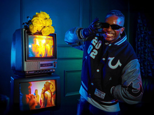 Man in a varsity jacket poses playfully in a dim room with vintage TVs stacked, displaying vibrant scenes. Yellow roses top one TV.