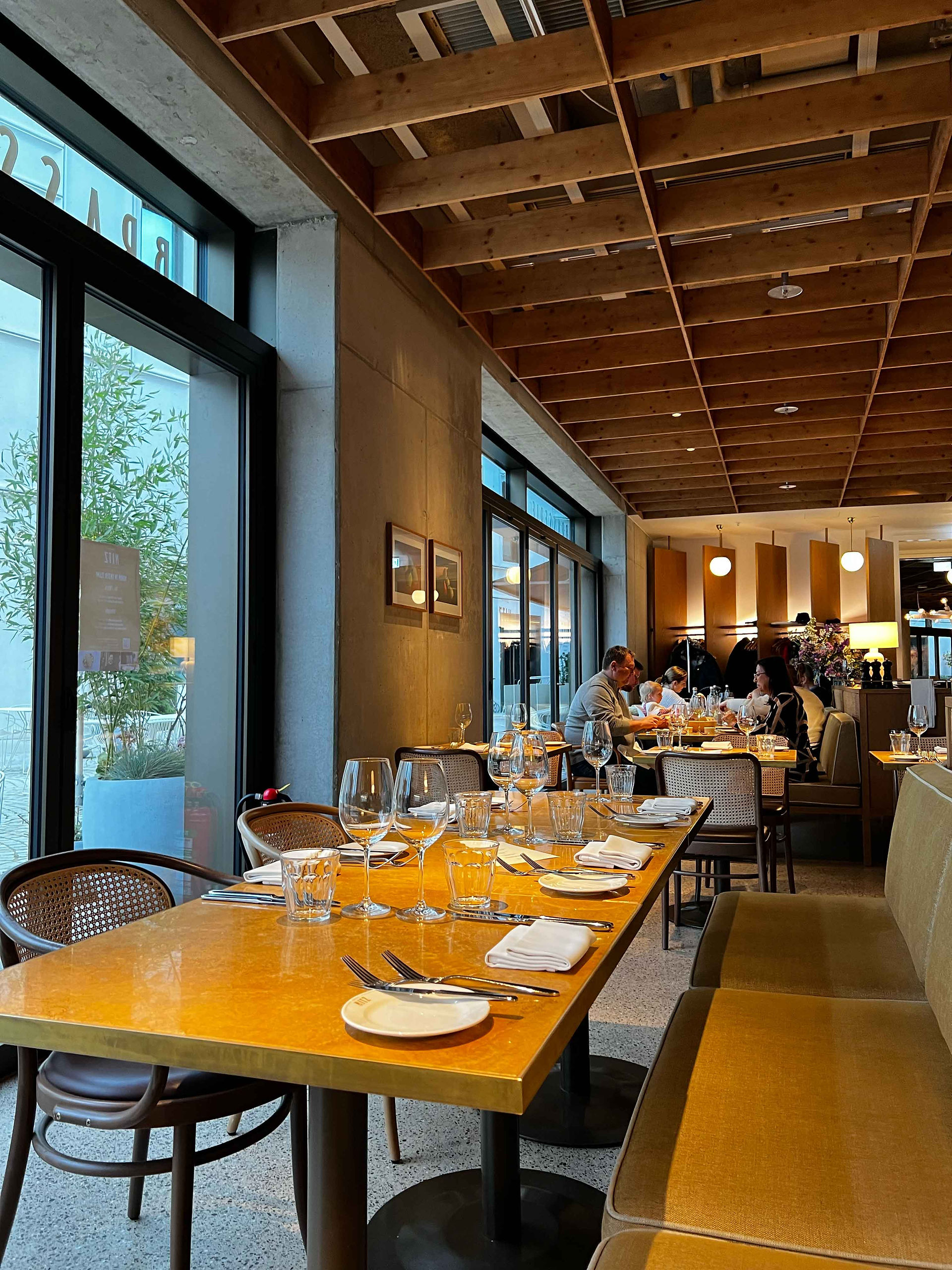 Eine gemütliche Speiseeinstellung in der Brasserie Nitz mit einem langen Tisch, der für eine Mahlzeit gedeckt ist, hölzerne Deckenbalken und große Fenster, die Tageslicht hereinlassen