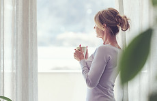 Mature woman relaxing at home and standing next to a window, she is looking away and drink