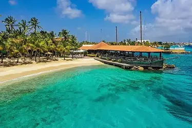 Luchtfoto van Harbour Village Beach Club op Bonaire met een wit zandstrand en palmbomen, helder turquoise water en een houten pier met oranje dak die uitsteekt in de baai