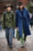 Two people walk on a cobblestone street. One wears a green jacket and striped pants, the other a blue coat and hat. Both appear stylish.