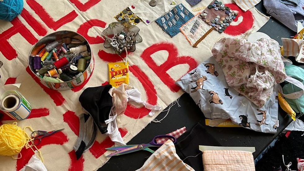 Crafting station with fabric scraps, scissors, thread, and decorative patches on a table. "HILLIES CLUB" painted in red on cloth.