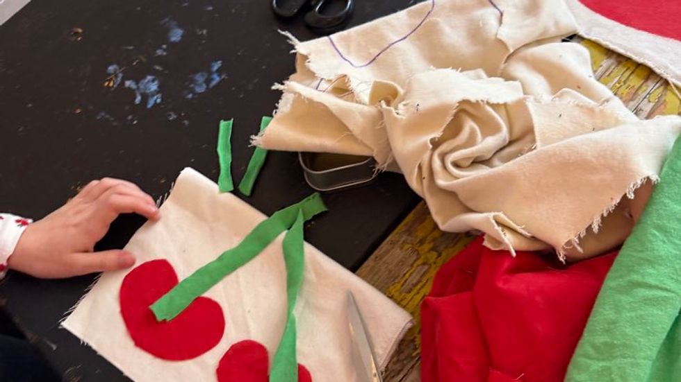 Child's hands crafting cherry design with red and green felt on white fabric. Scissors and other sewing materials on a dark table.