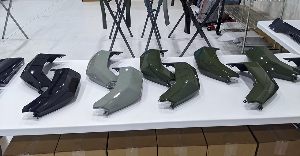 Glossy black, gray, and olive car body panels laid out on a white table in a workshop, with folding stands and boxes below.