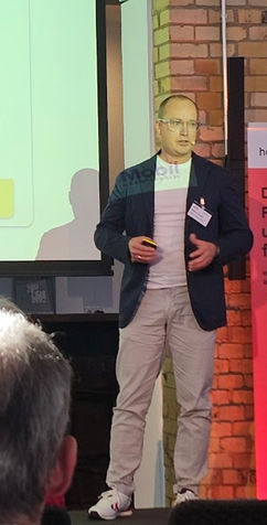Foto eines Mannes mit Brille, Sakko und hellem Shirt bei einer Präsentation auf einer Bühne im Innenbereich. Er hält ein Mikrofon oder Präsentationsgerät in der Hand und spricht vor Publikum. Die Aufnahme vermittelt einen professionellen, engagierten und souveränen Eindruck.