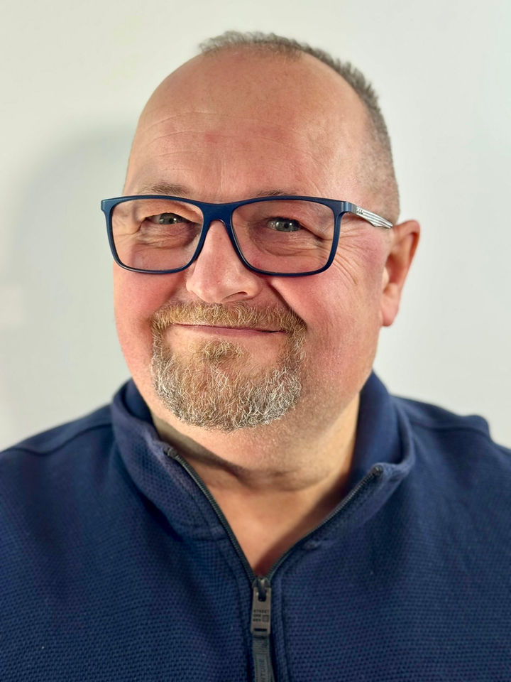 Portraitfoto eines lächelnden Mannes mit Brille und kurzem Bart vor hellem Hintergrund. Er trägt ein dunkles Oberteil und blickt freundlich leicht seitlich in die Kamera. Die Aufnahme vermittelt einen offenen, sympathischen und nahbaren Eindruck.