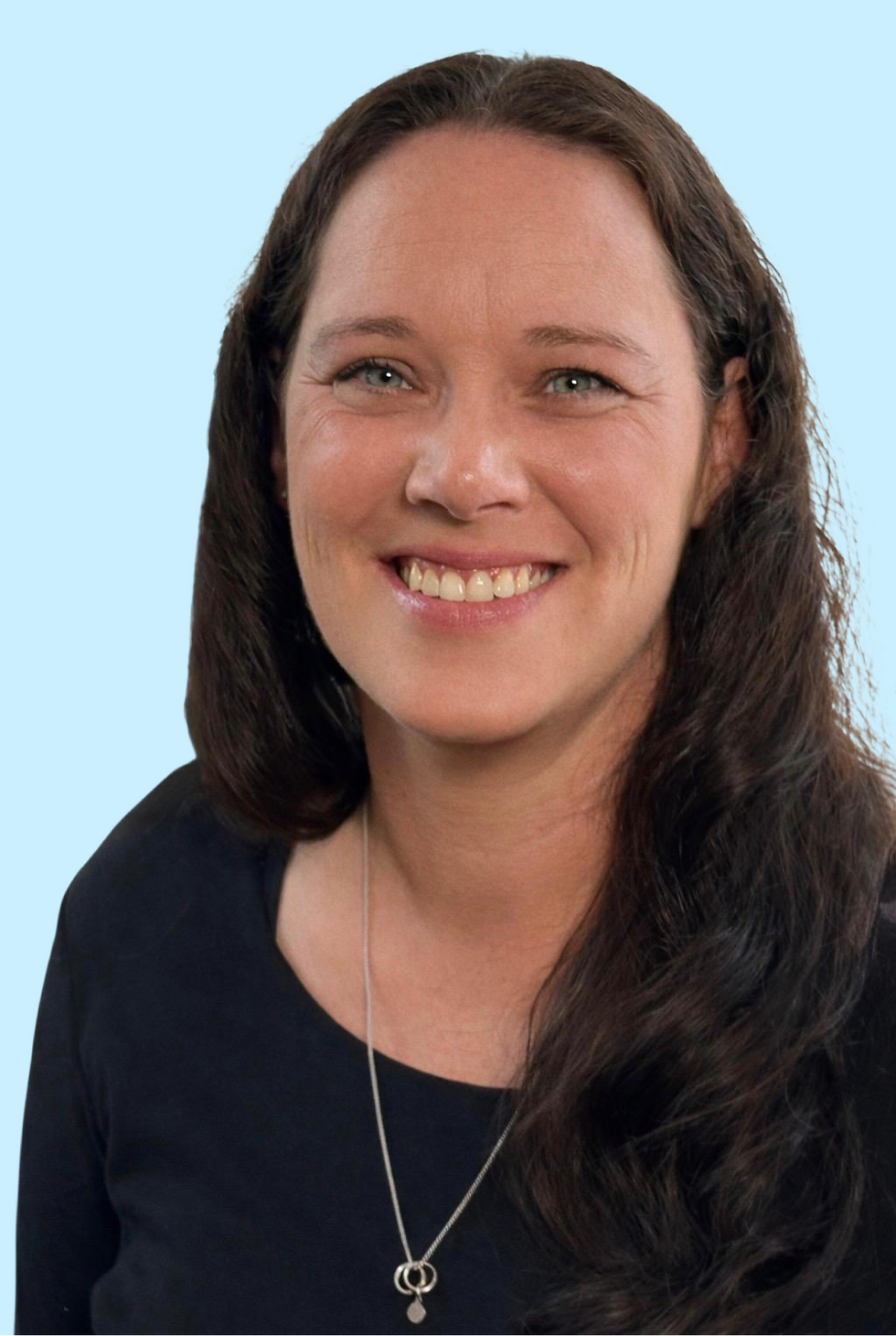Portraitfoto einer lächelnden Frau mit dunklem Haar vor hellem, einfarbigem Hintergrund. Sie trägt ein dunkles Oberteil und blickt freundlich in die Kamera. Die Aufnahme vermittelt einen offenen, sympathischen und nahbaren Eindruck.