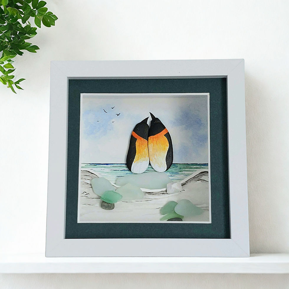 A white framed hand made 3D watercolour picture showing penguins made from pebbles perched on a piece of sea glass.