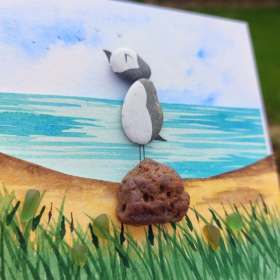 Thumbnail: Handmade framed puffin coastal art with painted pebbles, sea glass and a watercolour beach scene in a white frame with blue m