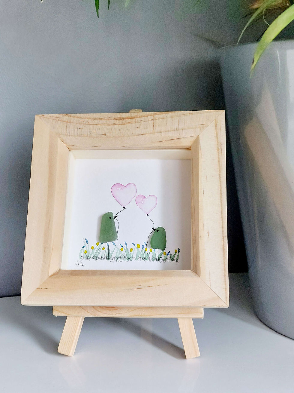 Thumbnail: A pine framed picture of two birds holding heart balloons in their beaks. The birds are made from sea glass.