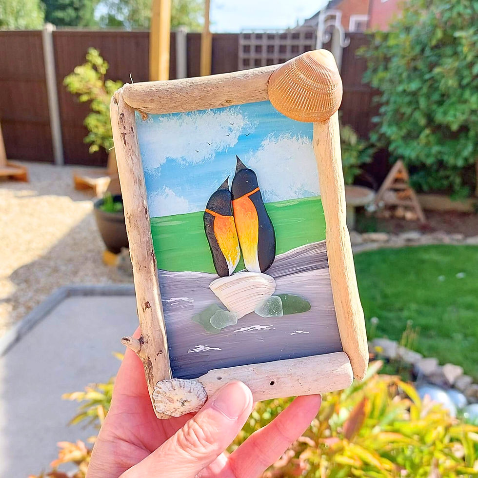 Handmade penguin couple pebble art featuring two hand-painted birds nestled together by the sea, framed with driftwood