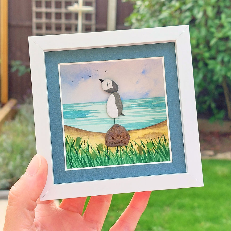 Handmade framed puffin coastal art with painted pebbles, sea glass and a watercolour beach scene in a white frame with blue m